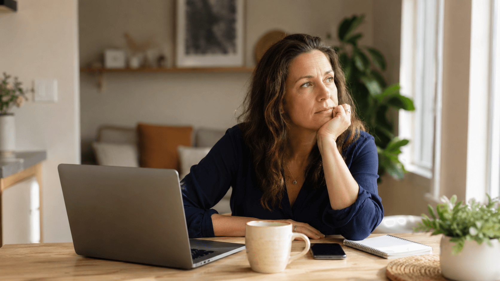 Woman sitting at a home table with a laptop, coffee, phone, and notebook, looking mentally stuck rather than lazy.