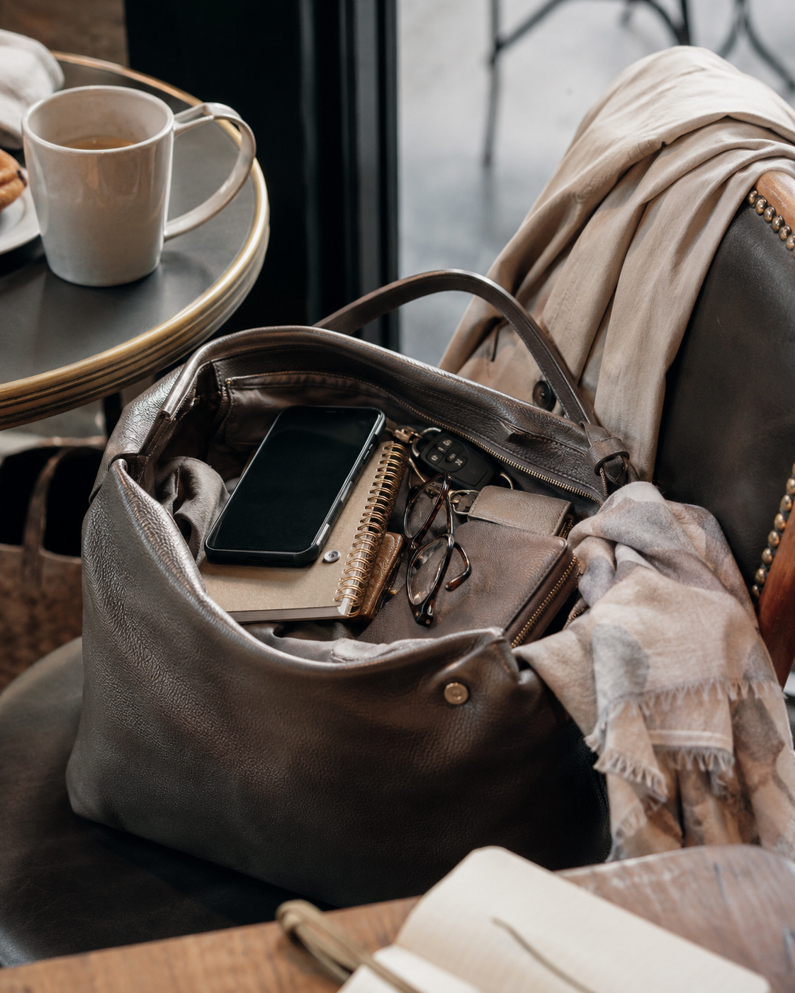 An open handbag with phone, keys, glasses, notebook and personal items at a café, representing people-pleasing, over-responsibility and always being available.